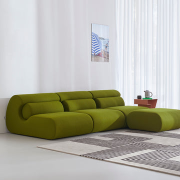 A modern living room highlights the green, modular curved design of the Ola Sofa by Objekte unserer Tage, featuring memory foam comfort and a matching ottoman. Light walls and floors complement a geometric rug, side table with teapot set, and Mediterranean-themed beach artwork.