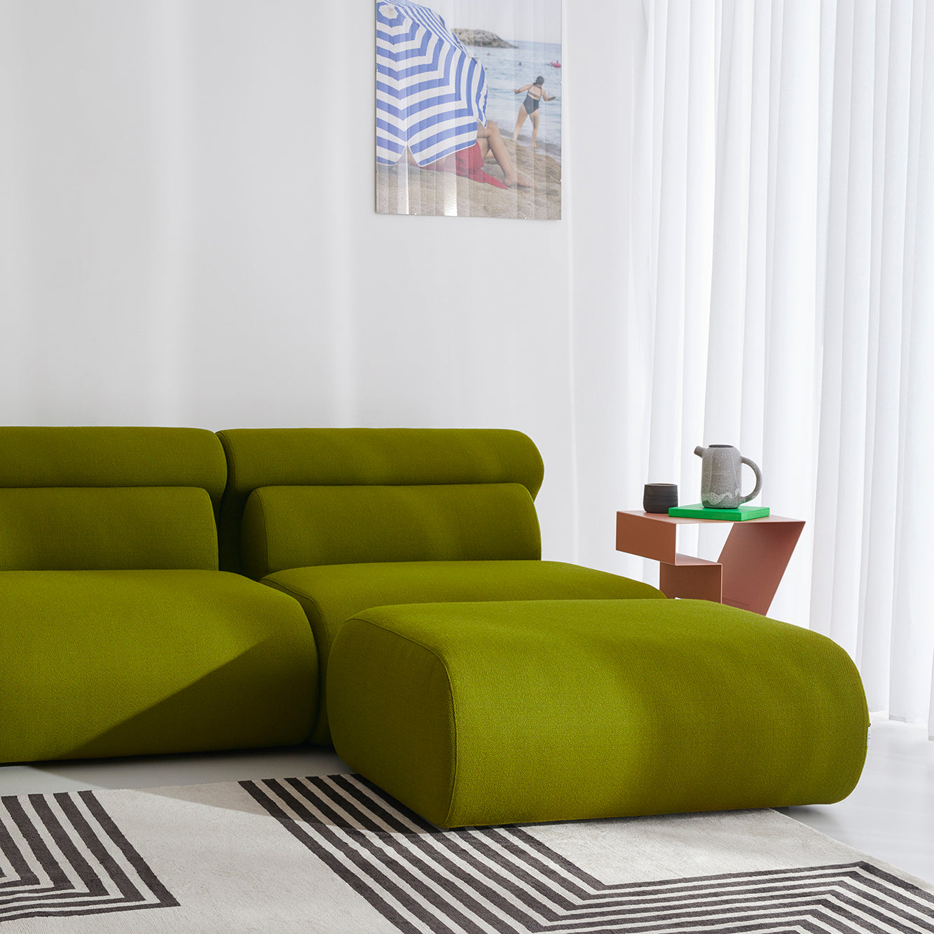 A modern living room showcases the Ola Pouf by Objekte unserer Tage in green, its modular design enhancing versatility on a geometric-patterned rug. A small angular side table with a teapot and cup complements the scene. Sheer white curtains frame the window, while a beach photo adds serenity.