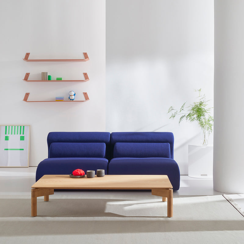 A modern living room showcases an Objekte unserer Tage Ola Sofa in calming blue, paired with a wooden coffee table and wall-mounted shelves for books and decor. Sunlight enhances the minimalist design with memory foam cushions and Mediterranean greenery touches.