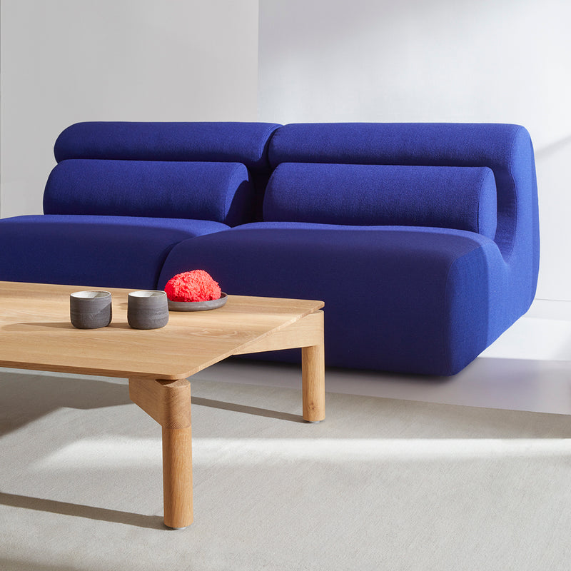 A modern living room features the Ola Sofa by Objekte unserer Tage, with bold blue, rounded seats and backrests. A light wooden coffee table with two small black cups and a red decorative piece complements the Mediterranean Sea-inspired decor for a minimalist and contemporary vibe.