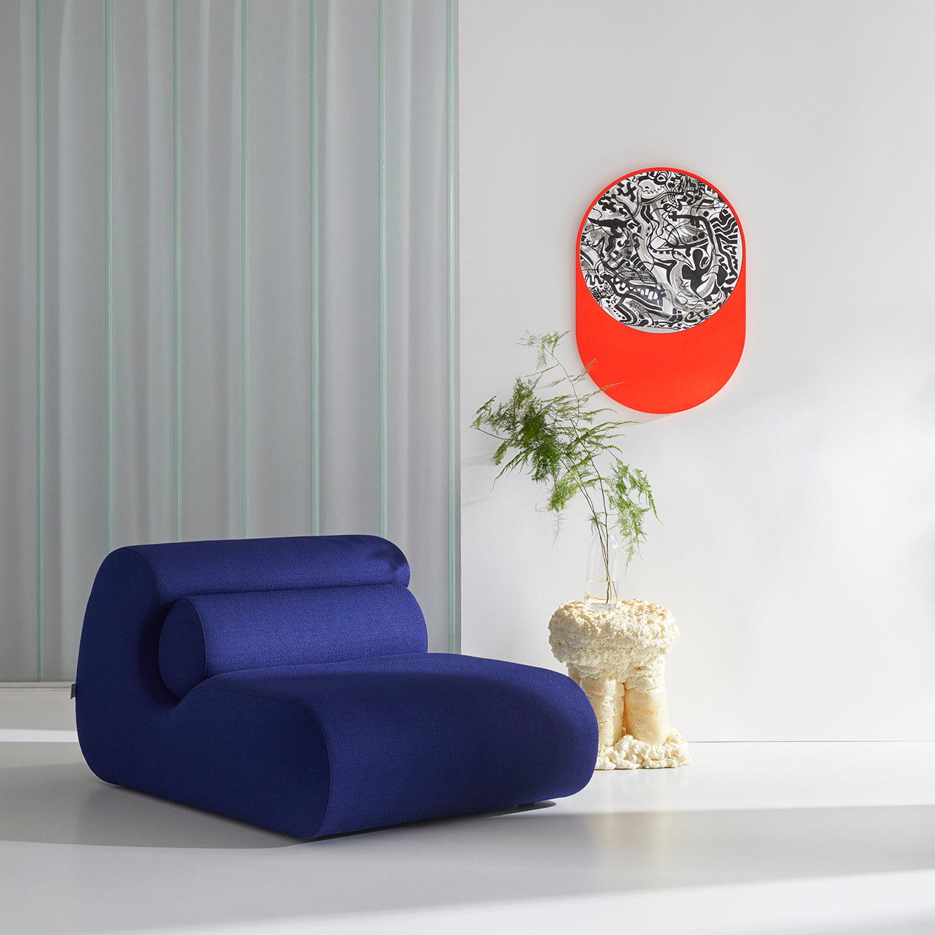 A modern room showcasing a blue, curvy Ola Sofa by Objekte unserer Tage, a sculptural end table with a small plant, abstract black and white artwork accented in bright orange on the wall. The minimalist decor is enhanced by light-colored walls and floors reminiscent of the Mediterranean Sea.