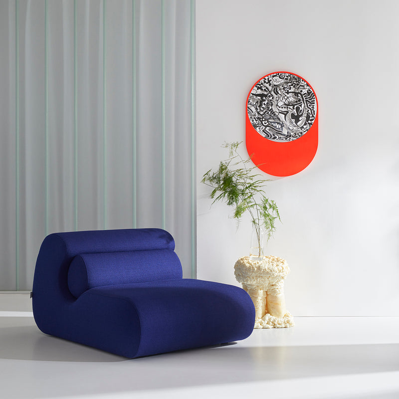 A modern room showcasing a blue, curvy Ola Sofa by Objekte unserer Tage, a sculptural end table with a small plant, abstract black and white artwork accented in bright orange on the wall. The minimalist decor is enhanced by light-colored walls and floors reminiscent of the Mediterranean Sea.