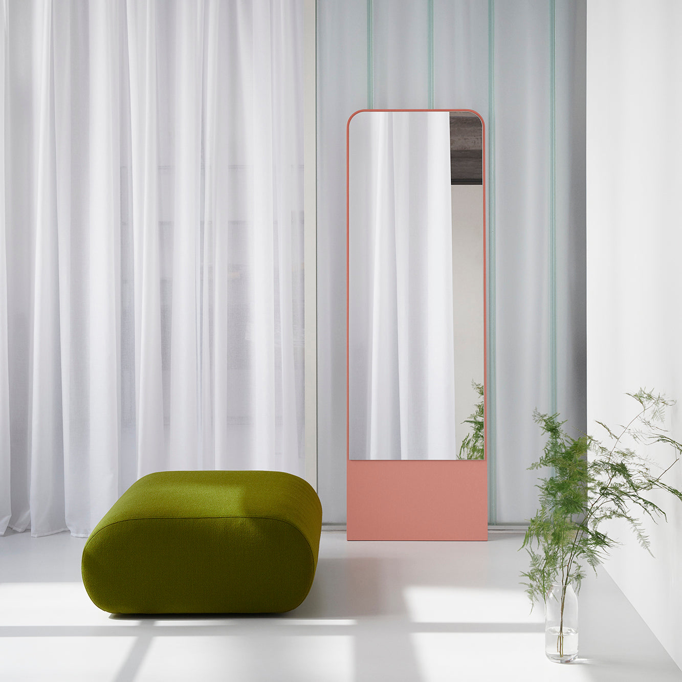 A modern interior features a tall, peach-framed rectangular mirror against a wall with white sheer curtains. The Ola Pouf, a green ottoman by Objekte unserer Tage with memory foam, complements the setting next to a small potted plant, creating a minimalist and serene atmosphere.