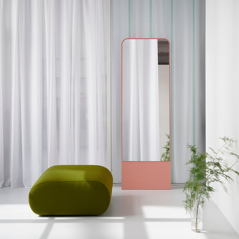 A modern interior features a tall, peach-framed rectangular mirror against a wall with white sheer curtains. The Ola Pouf, a green ottoman by Objekte unserer Tage with memory foam, complements the setting next to a small potted plant, creating a minimalist and serene atmosphere.