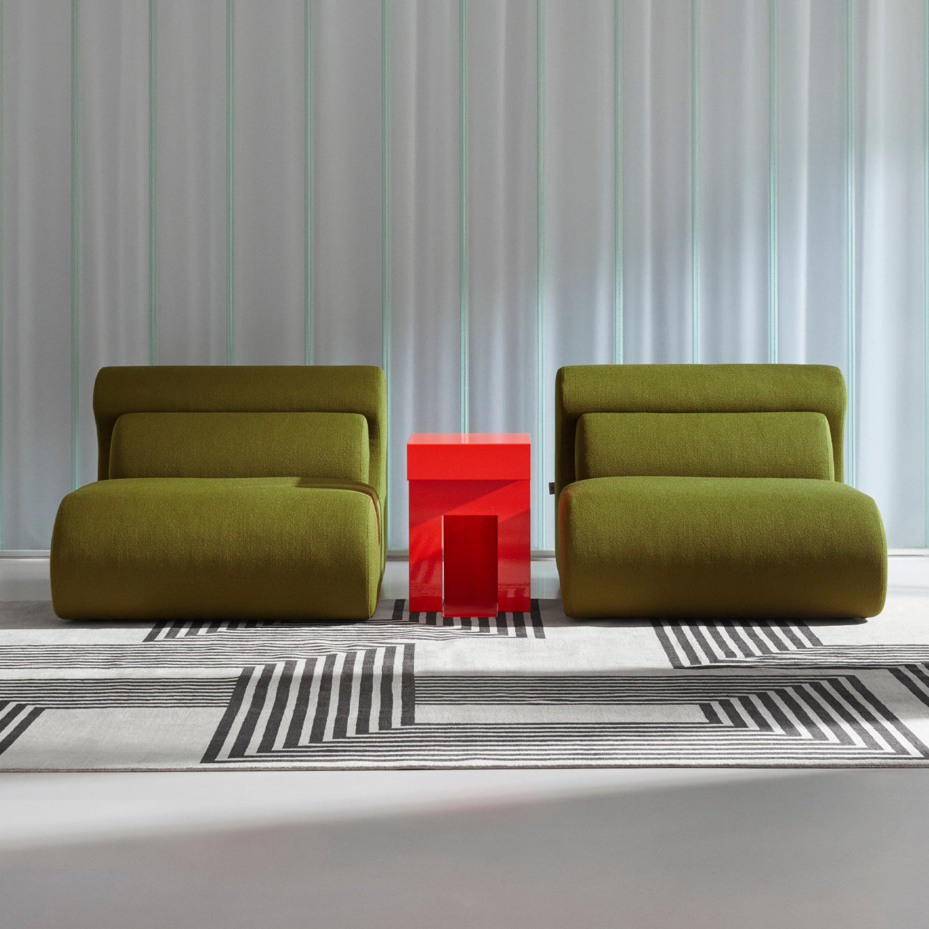Two Ola Sofas by Objekte unserer Tage, in modern green and designed with memory foam for ultimate comfort, sit on a patterned gray and white rug. A bright red table stands between them, against a backdrop of light, semi-transparent curtains for a minimalist contemporary setting.