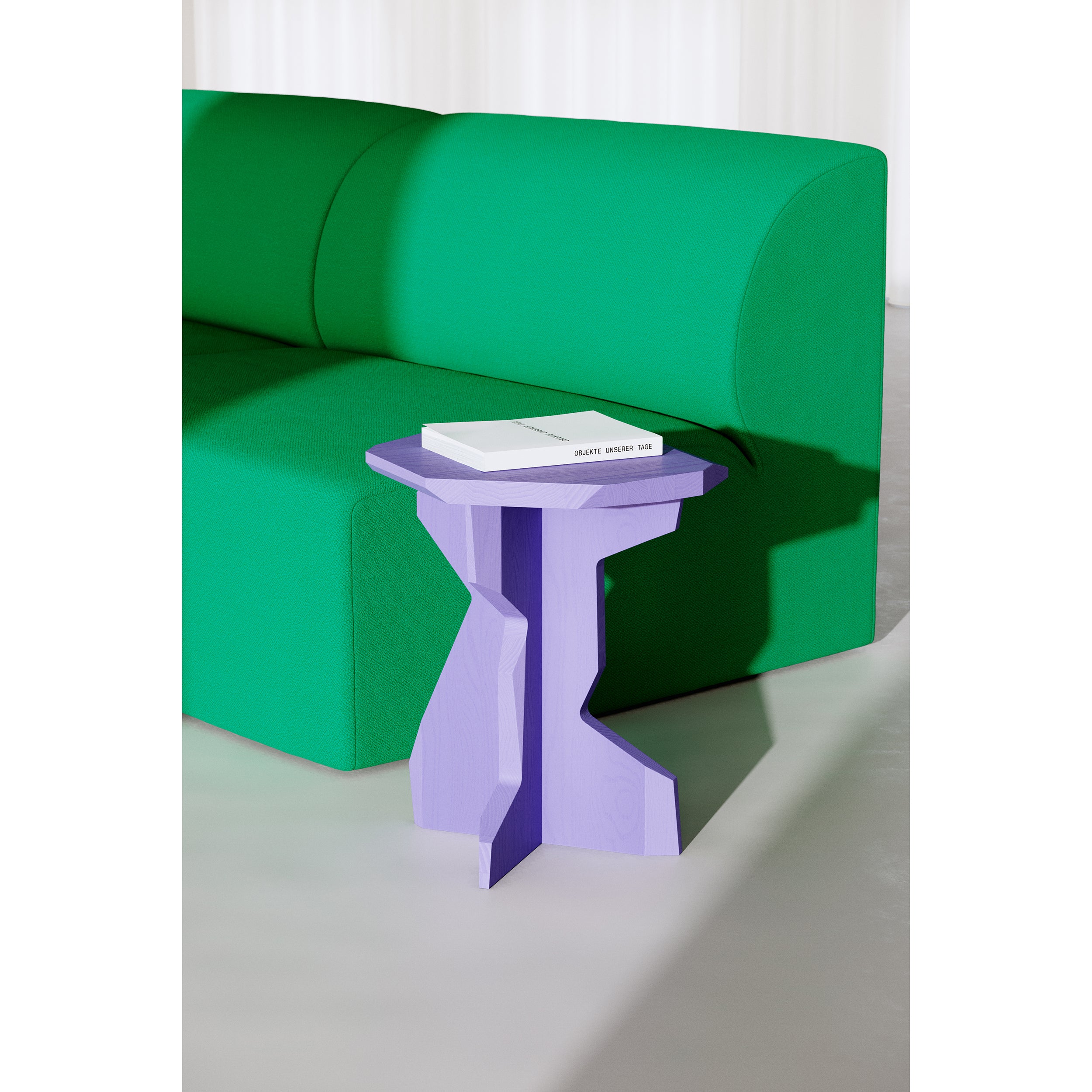 A modern living room includes a green sofa and the handcrafted Fels Sidetable by Objekte unserer Tage in purple geometric design. On it, a closed white book rests beside a natural wood stool. The well-lit room casts soft shadows, adding to its serene ambiance.