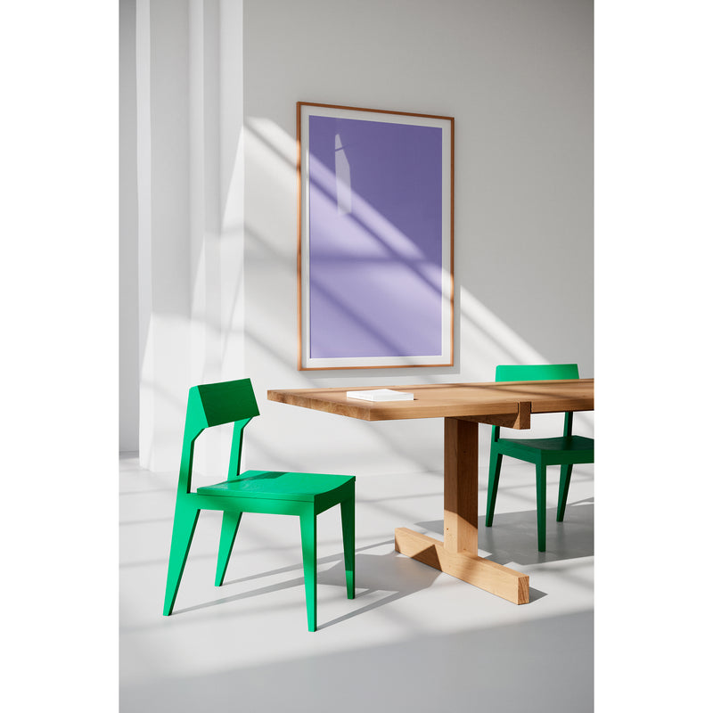 A modern interior includes two durable green Schulz Chairs by Objekte unserer Tage and a solid ash wood table. A purple and white abstract painting by SCHULZ enhances the wall, as sunlight creates serene shadows on the minimalist white floor and walls.