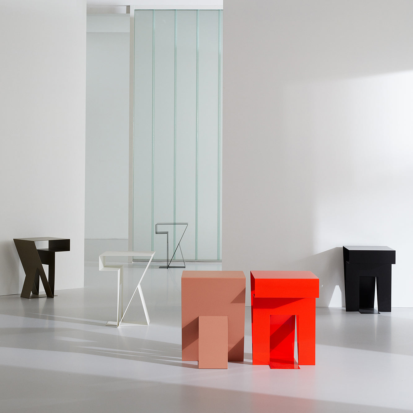 In a minimalist interior, five geometric stools in diverse colors and shapes stand out against a white and gray backdrop with a large frosted glass panel. The smooth floor and walls enhance the modern aesthetic, reminiscent of Objekte unserer Tages Neumann Sidetable design.