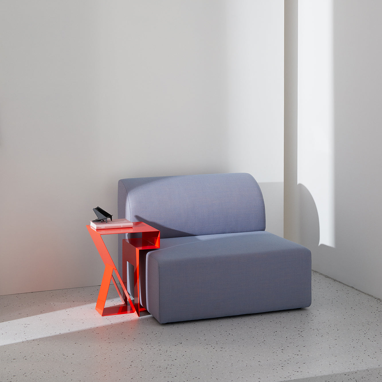 A minimalist room has a blue-gray sofa with a sleek, angular Neumann Sidetable by Objekte unserer Tage beside it. A small black object and a pink book rest on the table, reflecting its thoughtful design. Sunlight casts geometric shadows on the speckled floor and white walls.