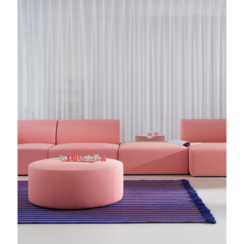A modern living room showcases a Weber Modular Sofa by Objekte unserer Tage in pink with a round ottoman. A blue and purple striped rug accents the floor, while a chess set sits on the ottoman. Sheer white curtains drape the windows, and an open book rests enticingly on the sofa.