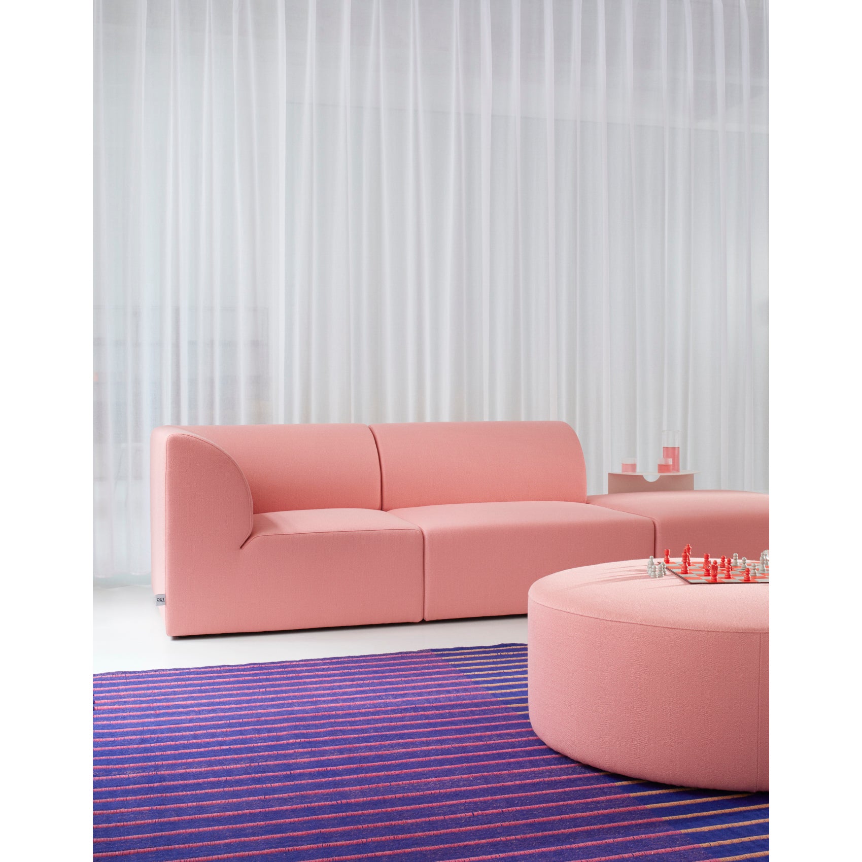 A modern living room exudes German elegance with Objekte unserer Tages pink Weber Modular Sofa and a matching round ottoman topped with a chess set. The floor features a blue and purple striped rug, while sheer white curtains create a soft, sustainable backdrop.