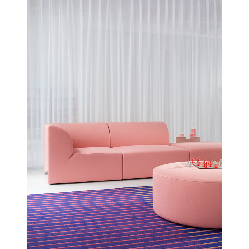 A modern living room showcases a pink WEBER modular sofa with a WEBER Pouf by Objekte unserer Tage, both in pink. A round ottoman holds small decor items, and a blue and pink striped rug completes the look. Sheer white curtains frame the window with a chic geometric basic shapes design.