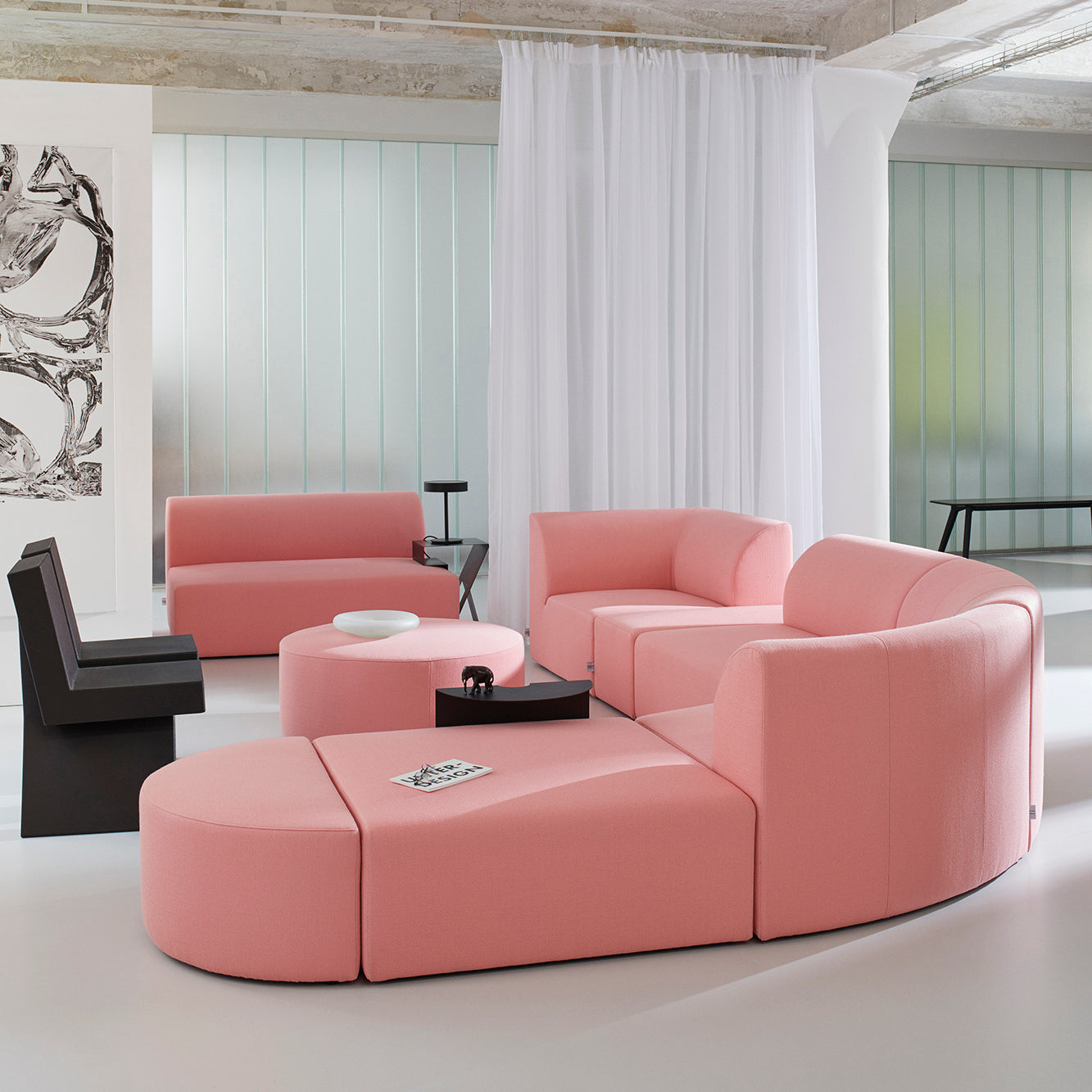 A modern living room showcases the Objekte unserer Tage Weber modular sofa in pink, arranged semi-circularly with a matching round ottoman. Nearby are a black chair and side table. The space exudes German sensuality through minimalist decor, large abstract art, and soft natural lighting.