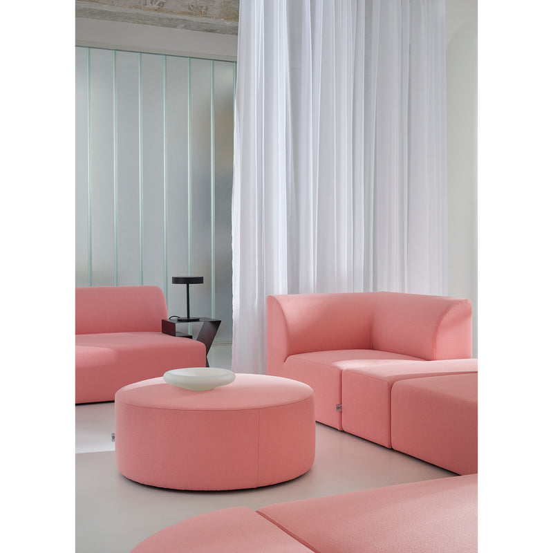 A modern living room showcases Weber modular sofas in pink from Objekte unserer Tage, with a matching round ottoman topped by a small white dish. Sheer white curtains and a black side table accentuate the bright, minimalist vibe with geometric shapes.