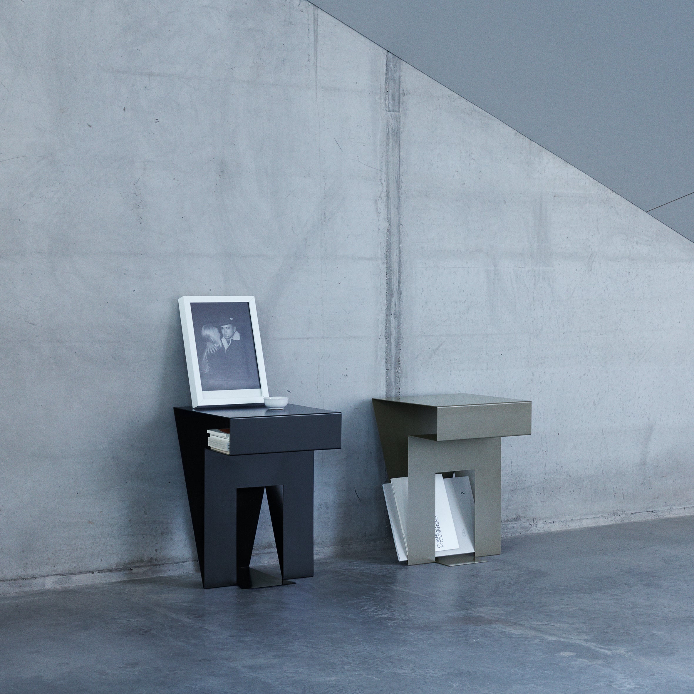 Two sleek Neumann Sidetables by Objekte unserer Tage stand by a concrete wall. The black one holds a photo, cup, and book; the gray stores magazines below. Diagonal lines from a staircase on the wall highlight elegant design.