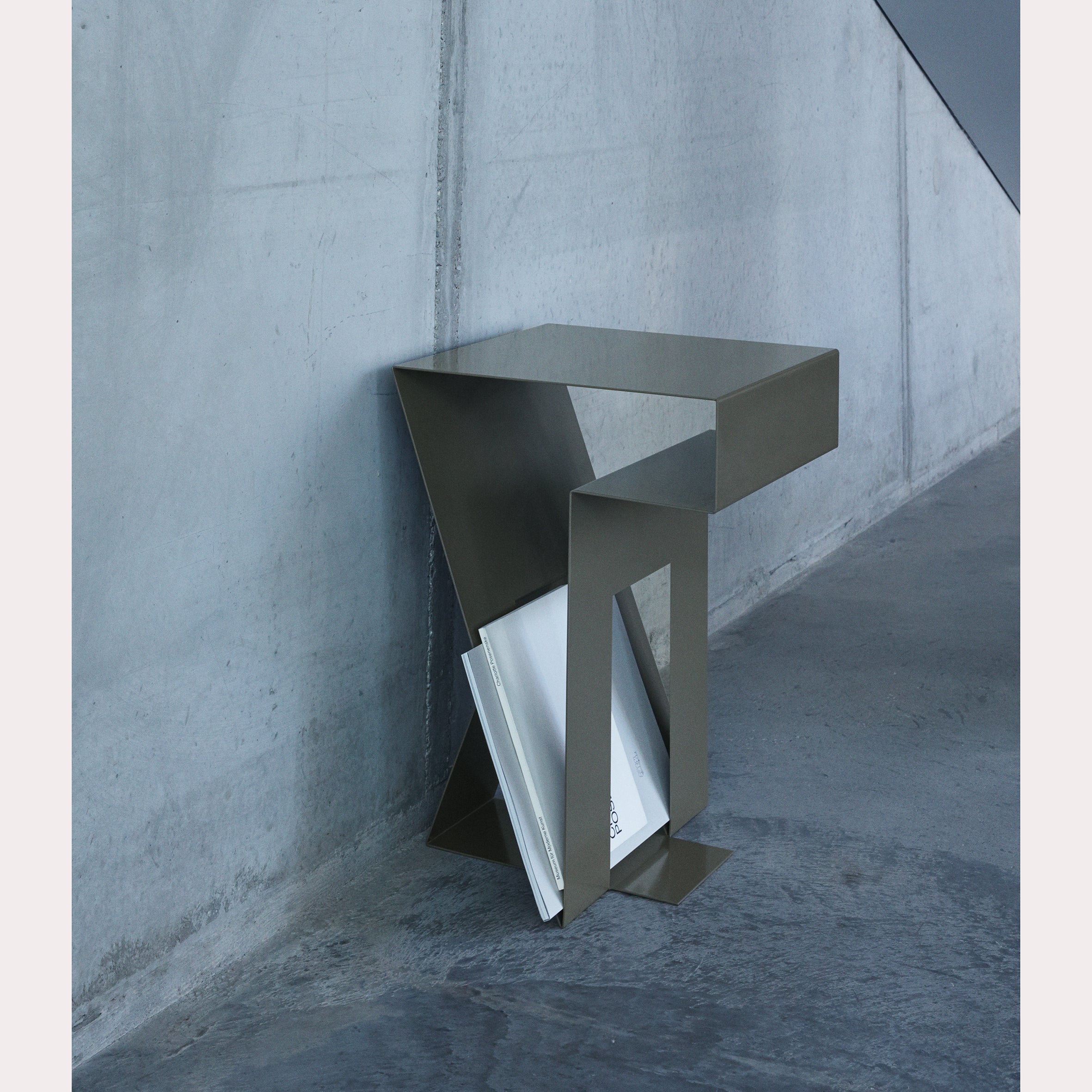 The Neumann Side Table by Objekte unserer Tage is a modern 3D design with an angular metal structure and built-in magazine holder. Its placement against a minimalist concrete wall on a polished floor showcases advanced design techniques in home decor.