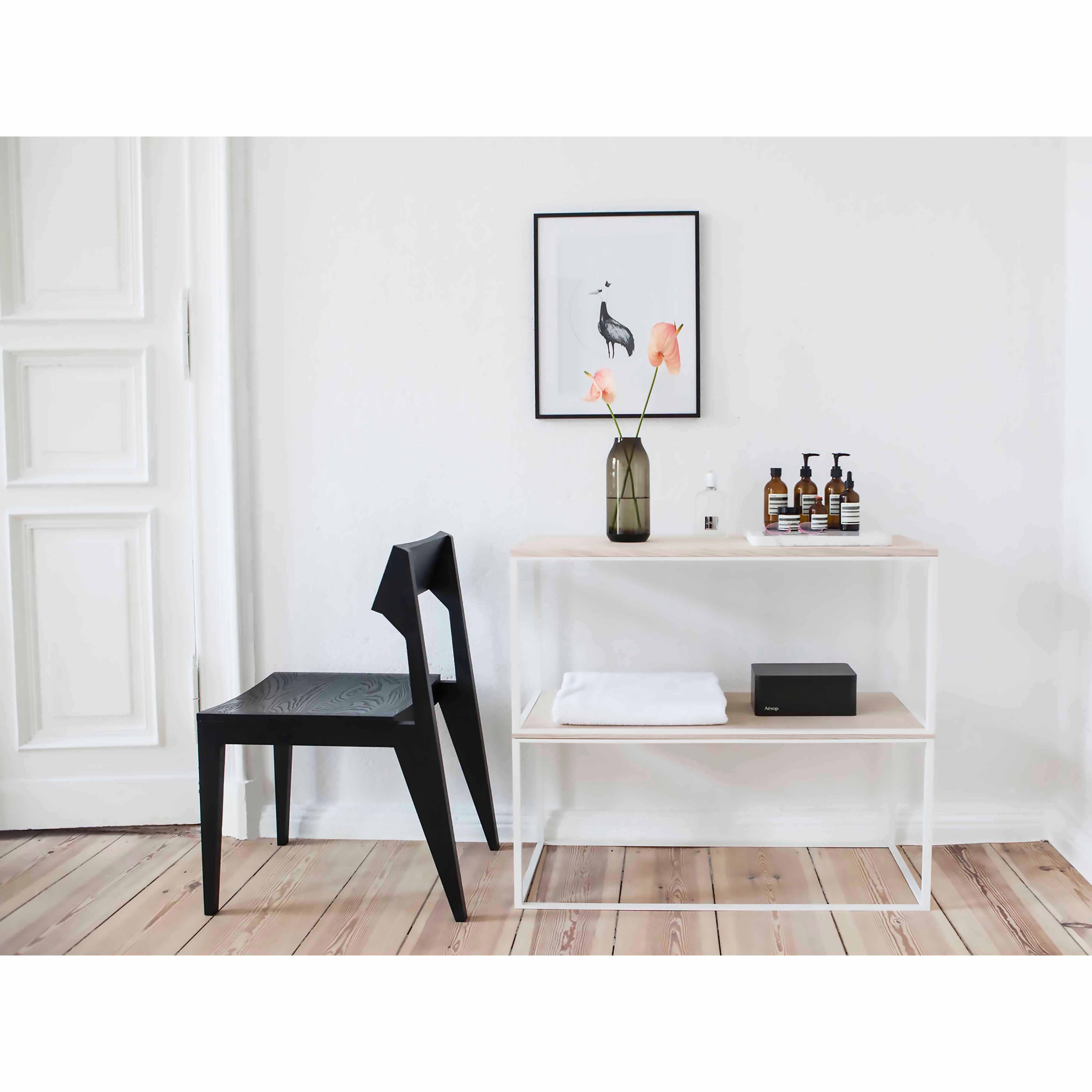 A minimalist room displays an Objekte unserer Tage Schulz Chair, celebrated for its durability, paired with a white shelving unit holding a vase with flowers and bottles. A floral artwork in a frame adds charm to the white wall, while the solid ash wood floor enhances the modern aesthetic.