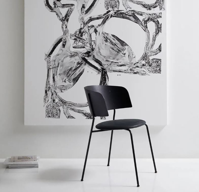 The Wagner Armchair by Objekte unserer Tage, featuring a minimalist design with powder-coated steel, provides comfort while standing elegantly on a glossy white floor. A large black-and-white abstract painting decorates the wall behind it, and a small stack of books sits to its left.