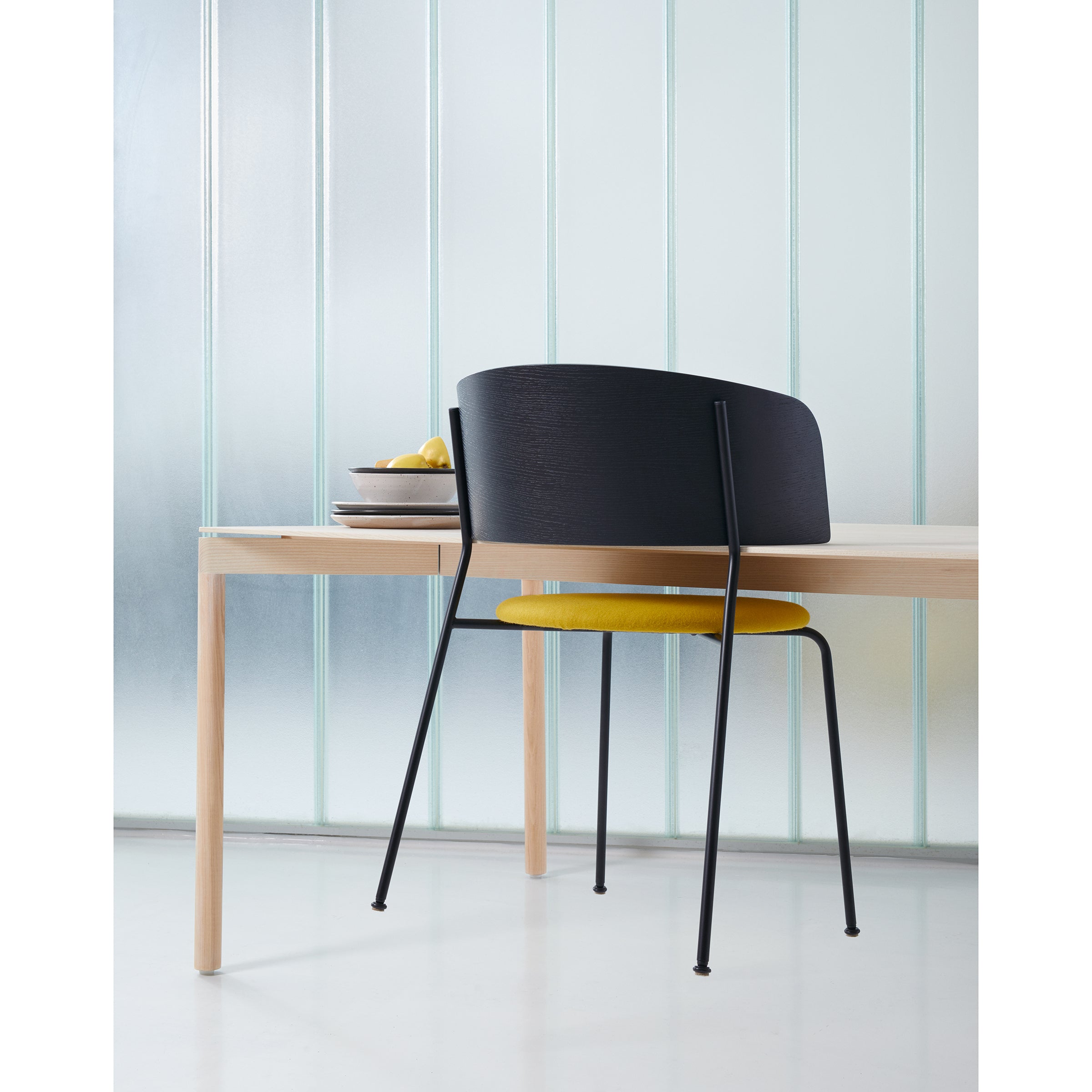 A minimalist dining setup includes a light wooden table and a stackable black chair with a yellow seat. Plates and lemons rest on the table, backed by a frosted glass wall with vertical lines. The WAGNER Armchair from Objekte unserer Tage, with its powder-coated steel frame, adds modern elegance.