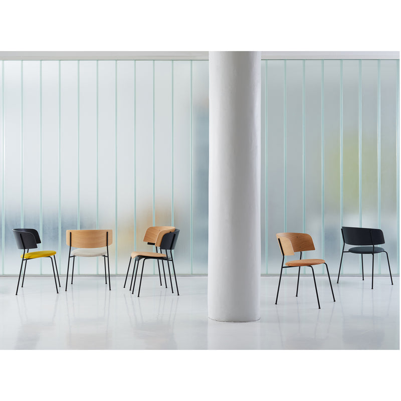 Seven modern, minimalist chairs with thin black legs, including the sleek Objekte unserer Tage Wagner Armchair and others with wooden and cushioned seats, are arranged near a frosted glass wall. Crafted from powder-coated steel, they enhance the rooms elegance around a central white pillar.