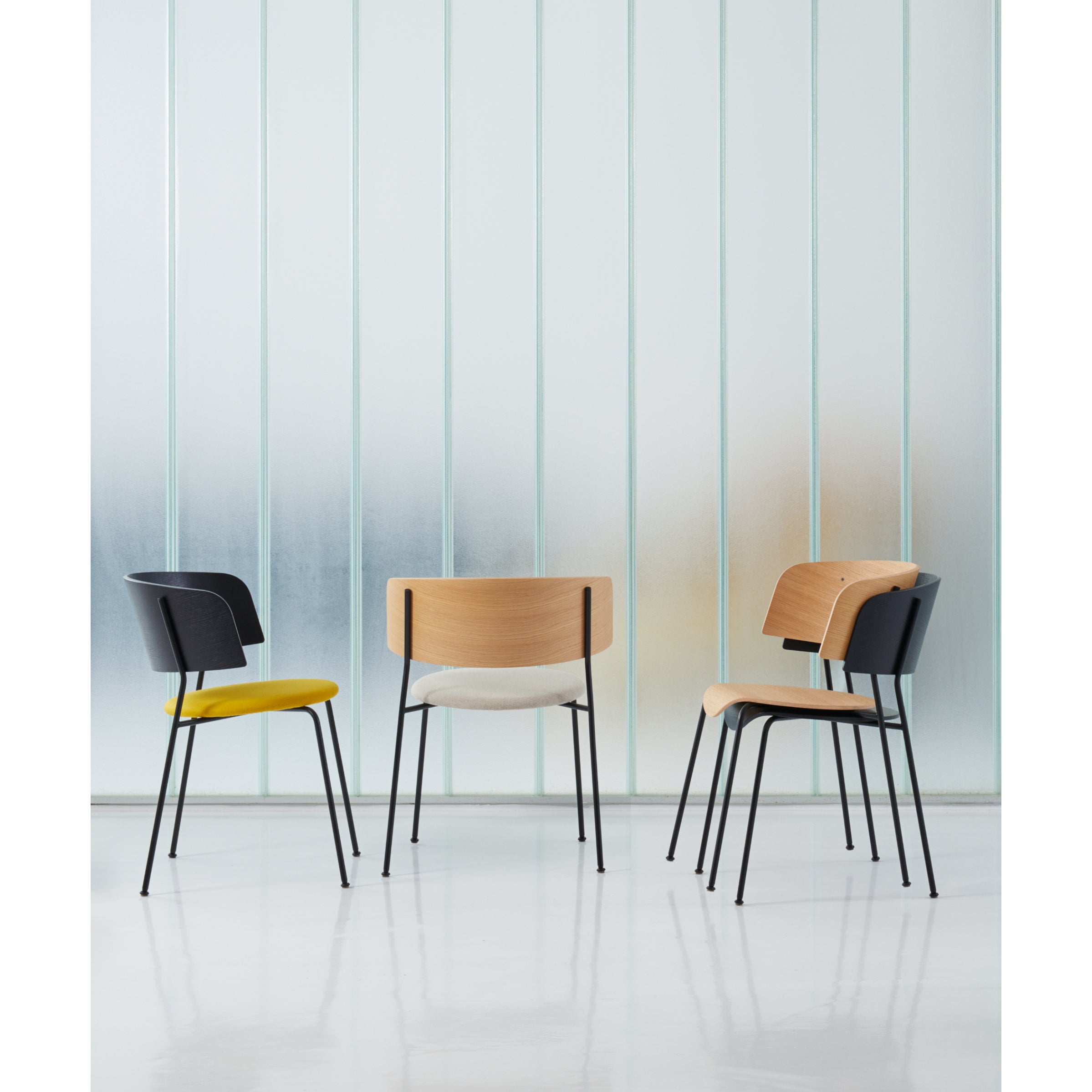 Three modern chairs with black metal legs are set in a minimalist space. Two have wooden backs; one has a black backrest. The seats are black, cream, and yellow. Included is the Wagner Armchair by Objekte unserer Tage. A frosted glass wall serves as the chic backdrop to this display.