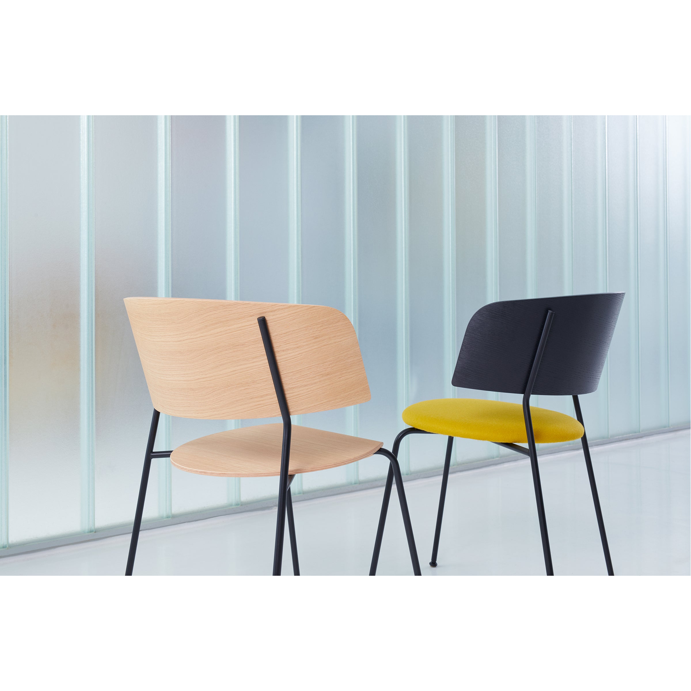 Two modern chairs with black powder-coated steel legs sit in front of a frosted glass wall. One has a light wooden back and seat, while the other, branded as the Objekte unserer Tage Wagner Armchair, features a dark back and bright yellow seat cushion.