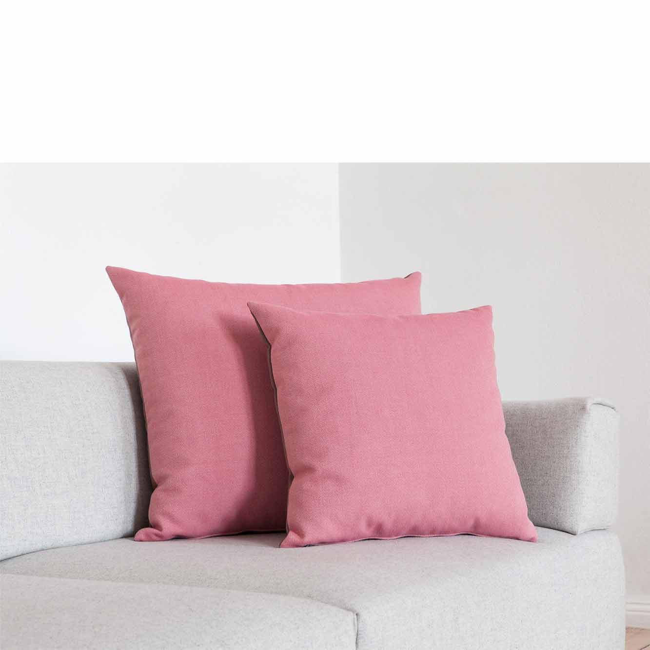 Two Weber Cushions in pink from Objekte unserer Tages collection are set on a light gray sofa in a minimalist room with white walls, adding a vibrant pop of color to the neutral-toned furniture.