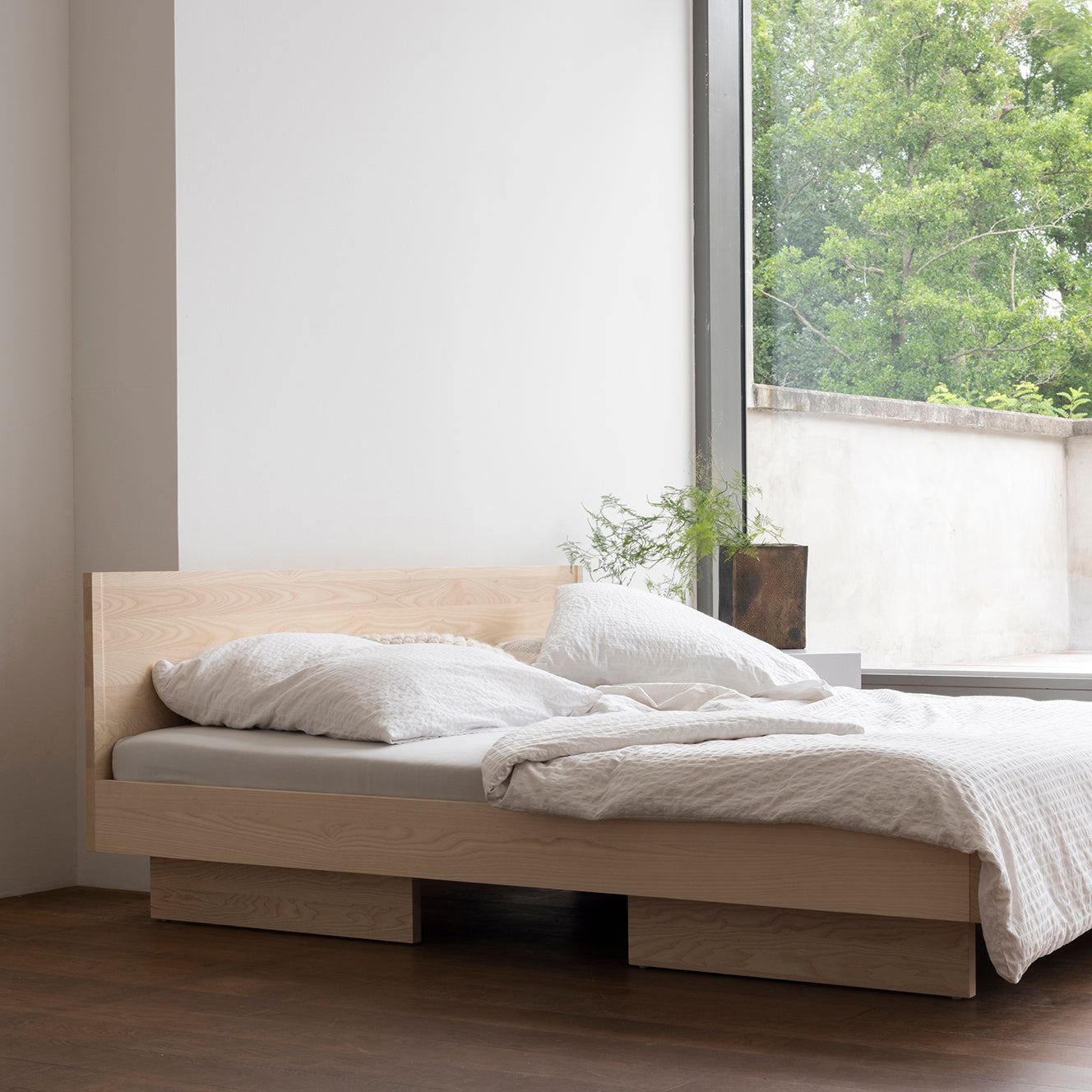 A minimalist bedroom features an Objekte unserer Tage Zians Bed with white bedding by a large window. The view of lush trees and a plant on the sill enhances the dreamy, cozy ambiance.