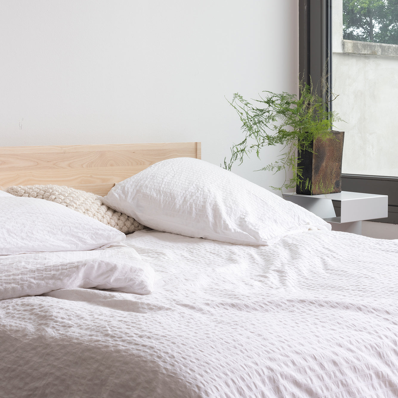 An Objekte unserer Tage Zians Bed in white bedding is positioned by a window. A potted green plant on a small white table enhances the dreamy coziness and natural sleeping ambiance of the minimalist bedroom.