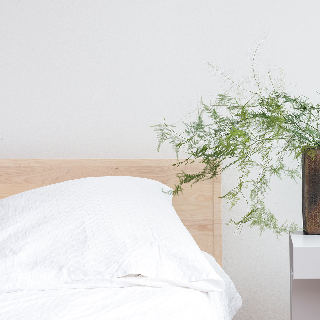 A minimalistic bedroom scene featuring the Zians Bed by Objekte unserer Tage with white linens exudes dreamy coziness. A side table holds a vase with lush, green foliage, and the plain white wall enhances the rooms simplicity and tranquility, crafting an atmosphere of natural sleeping bliss.