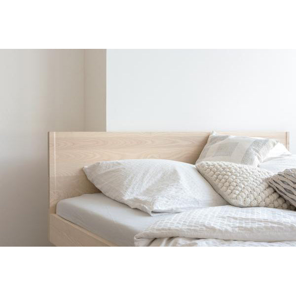 The Zians Bed by Objekte unserer Tage, featuring a light wooden headboard against a white wall, captures dreamy coziness. Dressed in white and gray bedding with a patterned comforter and textured pillows, it ensures natural sleeping comfort.