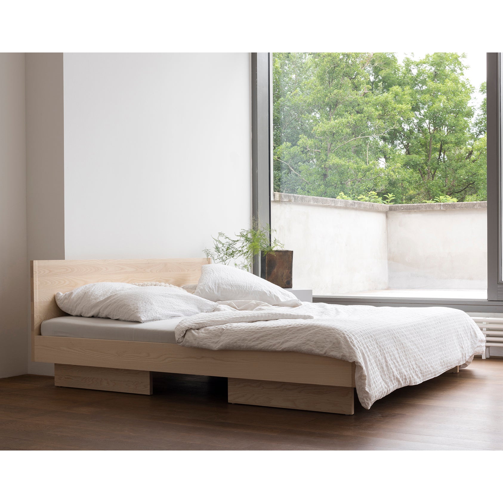 The minimalist bedroom exudes dreamy coziness with its Objekte unserer Tage Zians Bed, adorned in white bedding. A large window reveals green trees for natural comfort, complemented by a small plant and dark wooden flooring, creating a tranquil space.