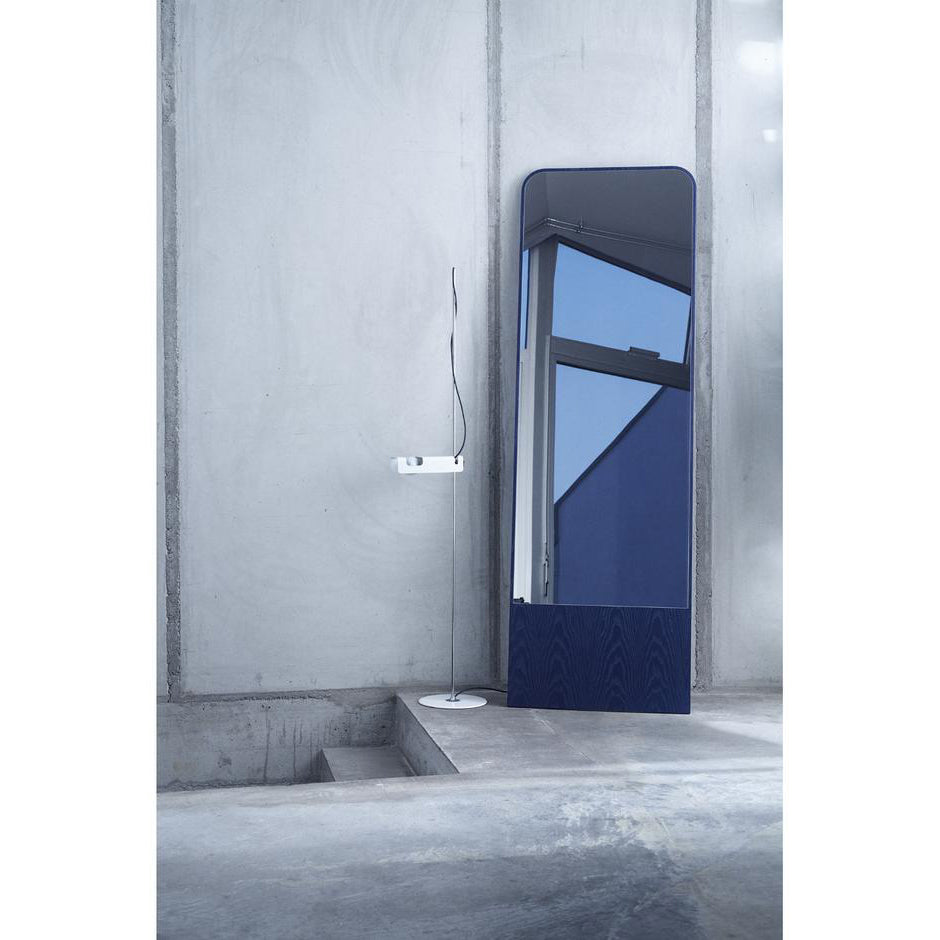 The Friedrich Mirror by Objekte unserer Tage stands against a concrete wall, exemplifying minimalist design. Its large, tall frame reflects a window with a clear blue sky. A slim floor lamp complements the mirror on the minimalist concrete staircase in this contemporary living space.