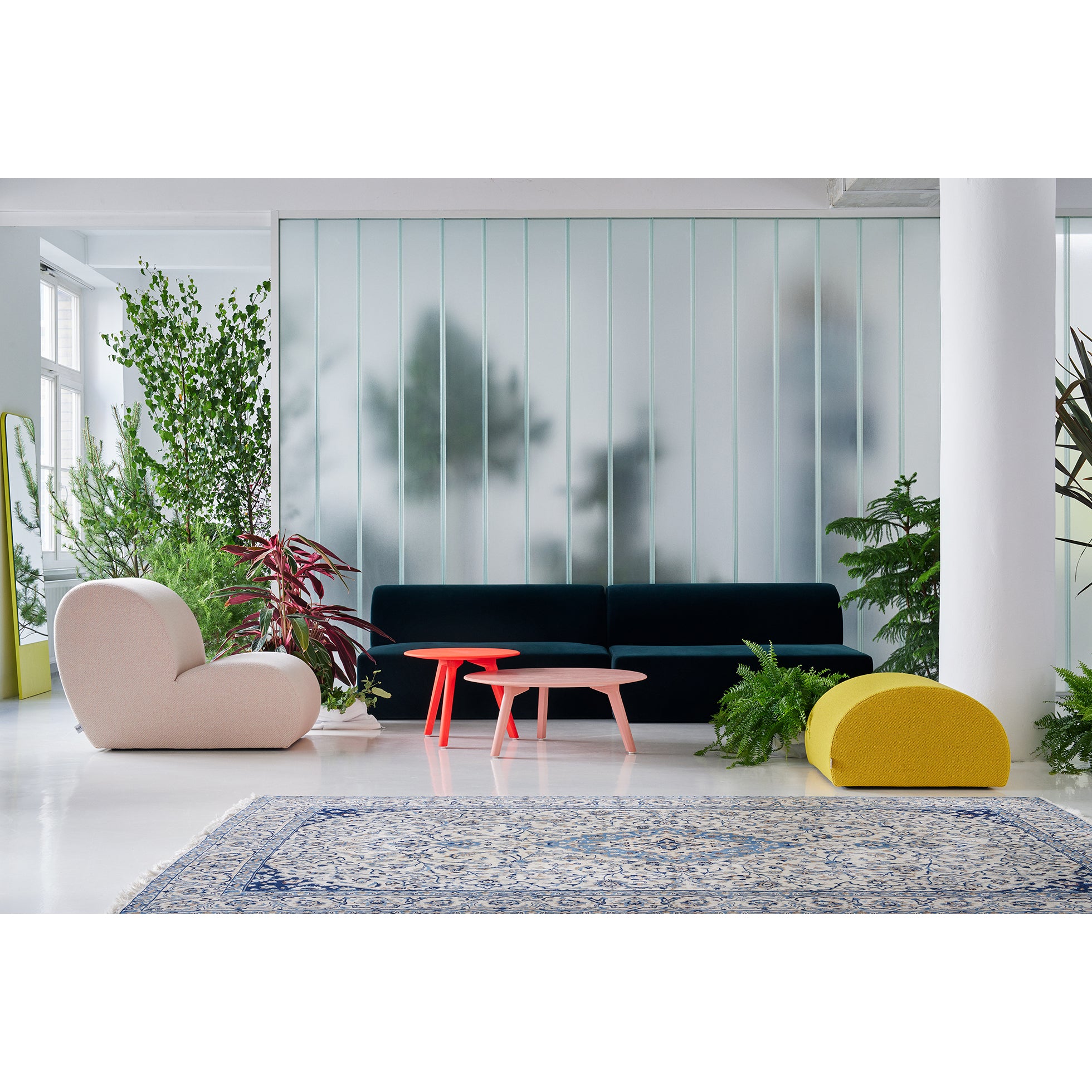 A modern living room features versatile upholstered furniture with a dark green sofa, the Levi Lounge Chair by Objekte unserer Tage in cream, and a yellow seat on a white floor. Two red coffee tables sit before the sofa, plants are scattered around, and frosted glass panels provide an elegant backdrop.