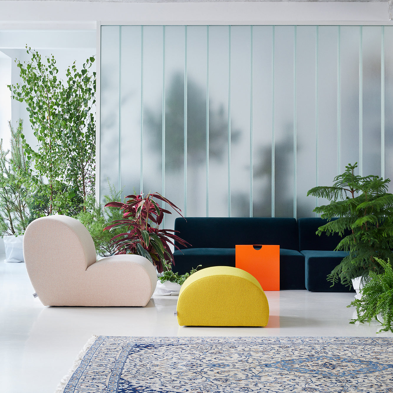 A modern living room features versatile upholstered furniture, including an Objekte unserer Tage Levi Lounge Chair, a yellow ottoman, a dark teal sofa, and a bright orange cube table. Lush green plants add serenity to the space, complemented by a frosted glass wall and patterned blue rug.