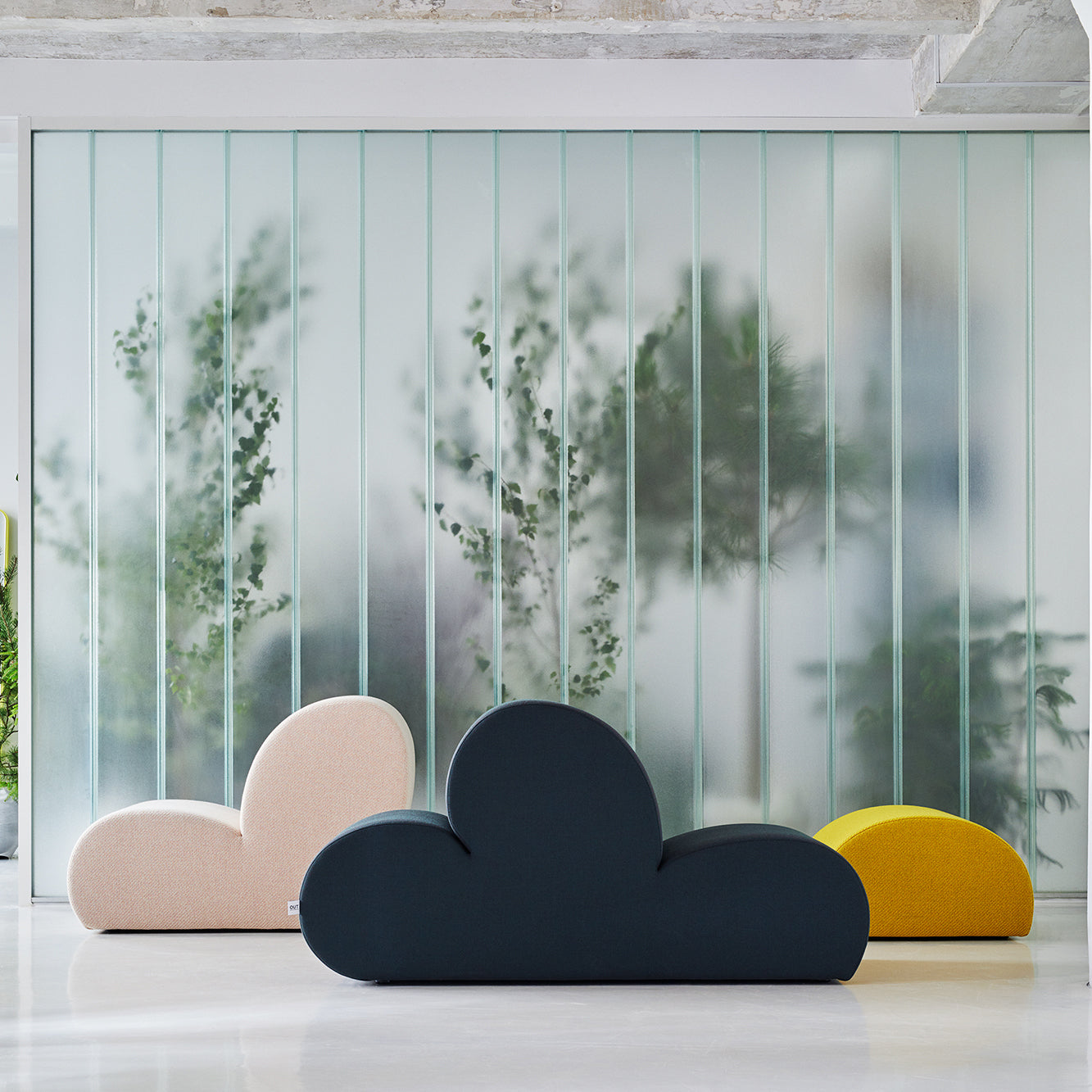 Three cloud-shaped Levi Lounge Chairs by Objekte unserer Tage in pastel pink, dark blue, and yellow adorn a glossy floor. A frosted glass wall with blurry green foliage is visible behind them, showcasing the handcrafted upholsterys minimalist and modern aesthetic.