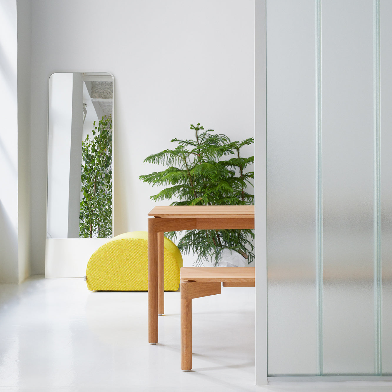 A minimalist room with a wooden table and bench features a large plant next to a full-length mirror that reflects greenery. A bright yellow Levi Pouf from Objekte unserer Tage is set against a white wall, while a frosted glass panel on the right enhances the airiness of the space.