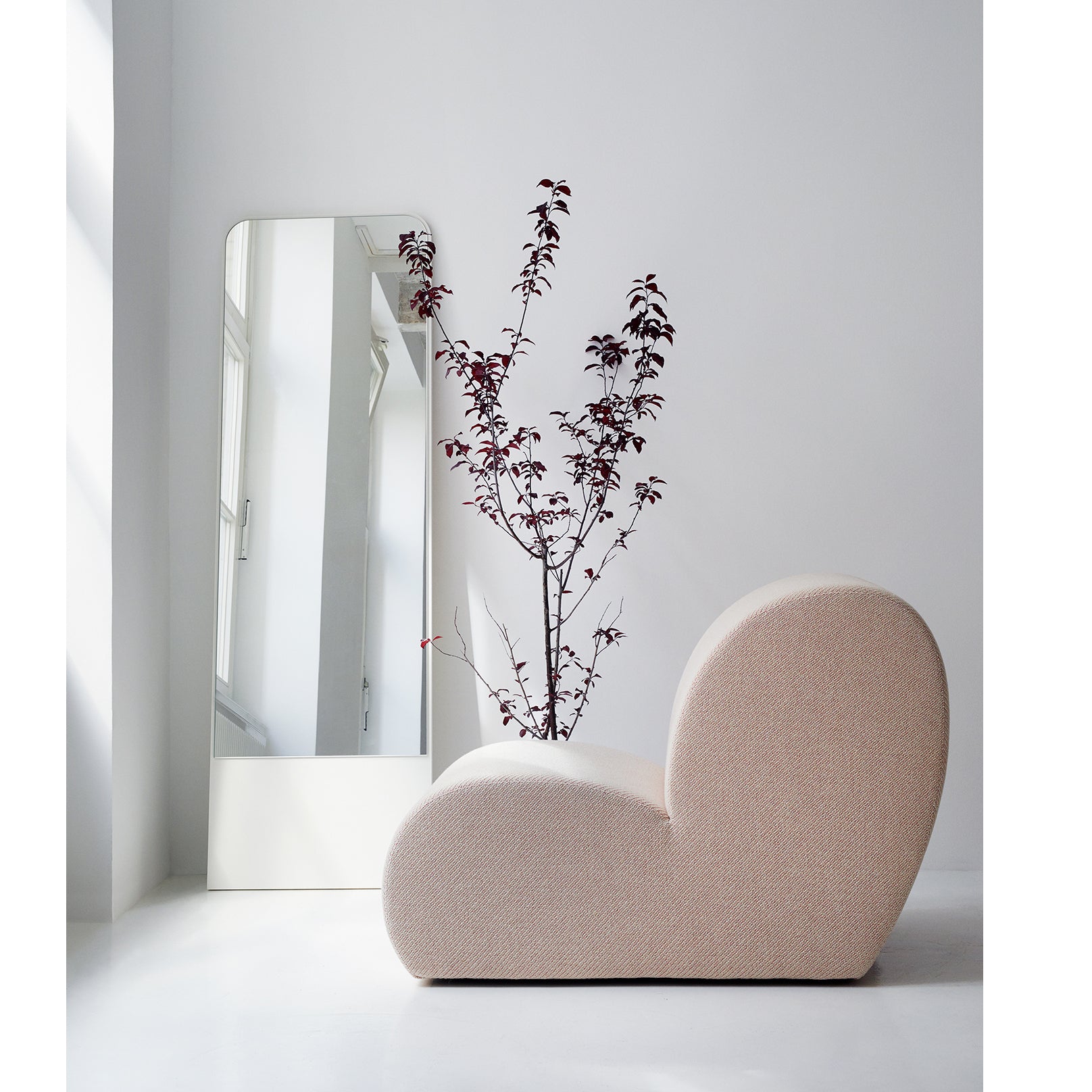 A minimalist room features the Levi Lounge Chair by Objekte unserer Tage, a modern beige chair with a curvy design. A tall mirror stands behind a slender plant with dark purple leaves against a white wall. Natural light from the left highlights its handcrafted upholstery.