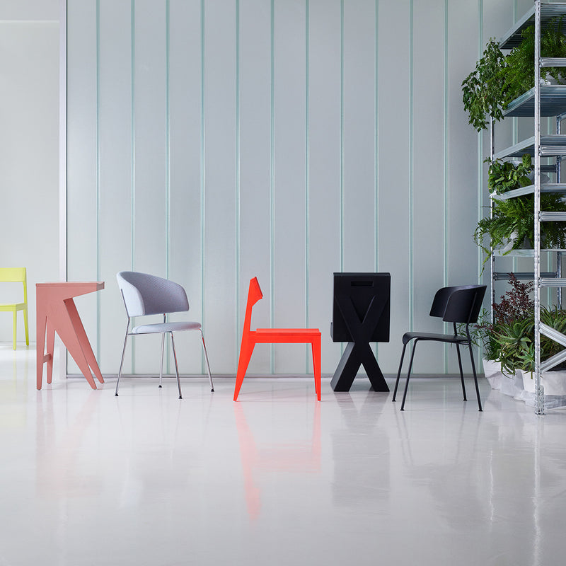 Five modern chairs, including the Objekte unserer Tage Wagner Dining Chair renowned for comfort, are displayed in diverse colors and styles on a glossy white floor. A shelving unit with green plants adorns the right side against a serene light green vertically lined wall backdrop.