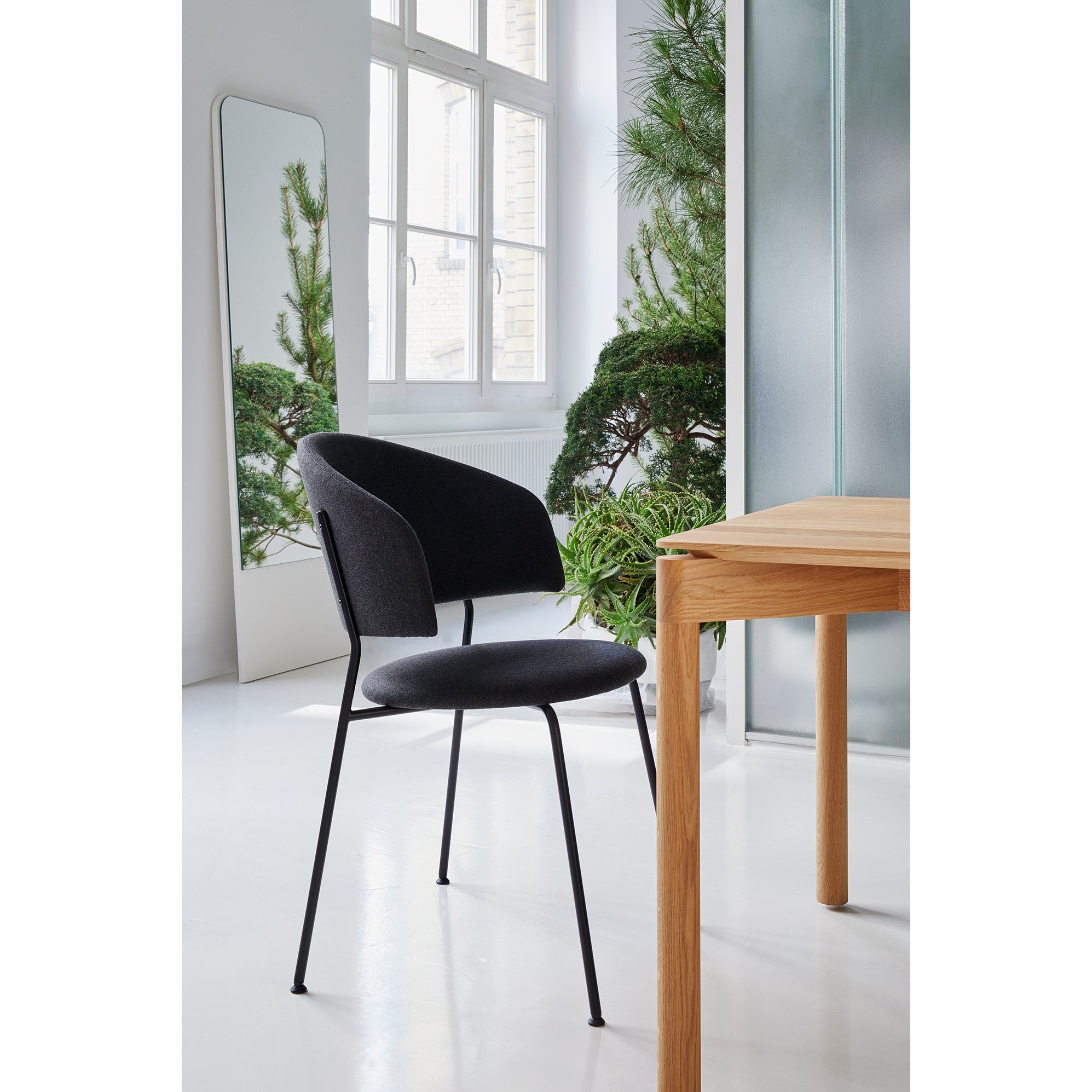 A modern dining area showcases the Wedekind Table by Objekte unserer Tage, made from sustainable oak, paired with a sleek black chair. A large mirror reflects green plants, enriching the bright, minimalist design. White walls and a window bathe the space in natural light.