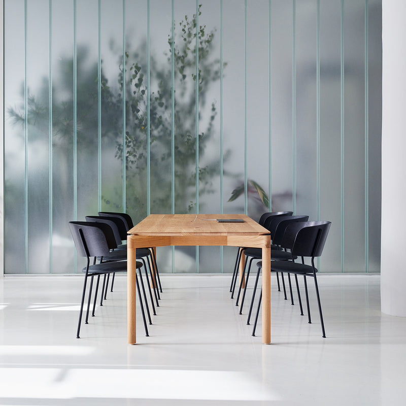 A minimalist conference room features an Objekte unserer Tage Wedekind Table crafted from sustainable oak, surrounded by black chairs. Frosted glass panels reveal plant silhouettes, all enhancing the rooms modern design and fine craftsmanship.