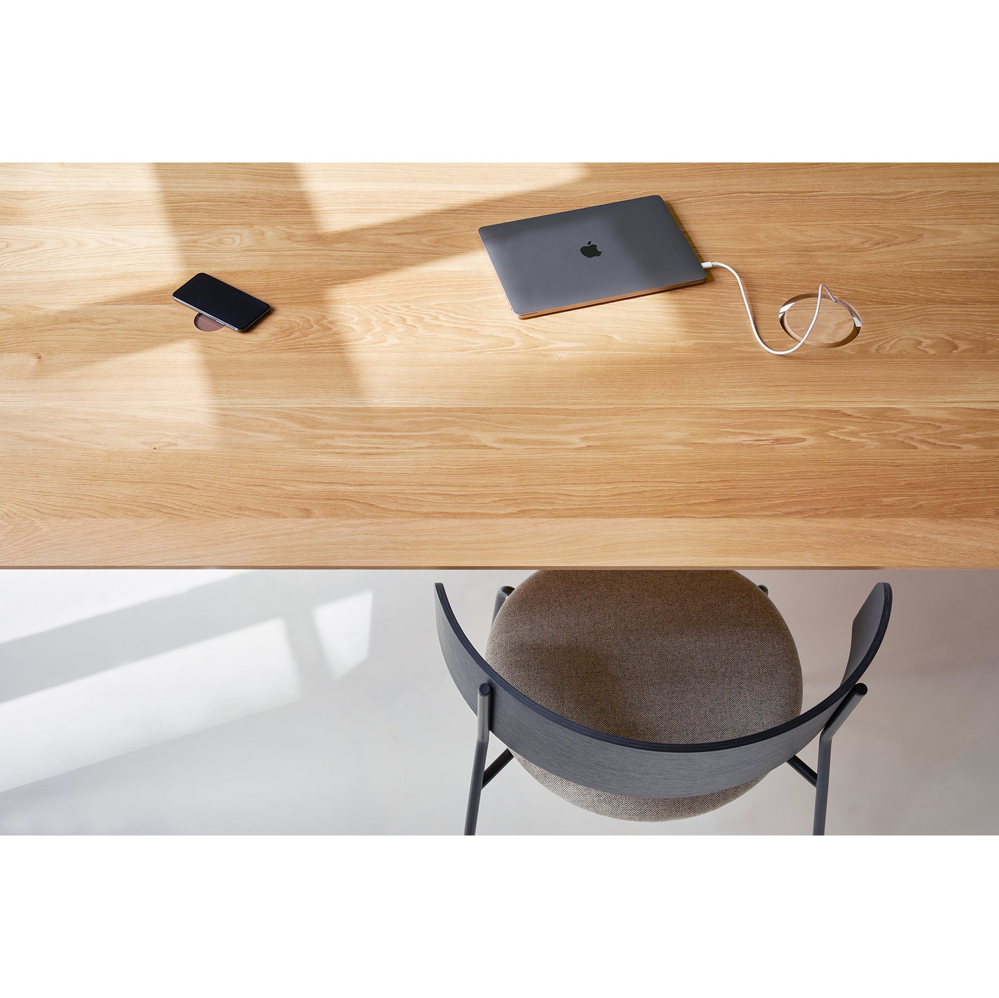 A Wedekind Table by Objekte unserer Tage, crafted from sustainable oak, holds a smartphone and charging laptop. A gray chair with a curved backrest is visible at the bottom, as shadows and natural light dance across the scene.