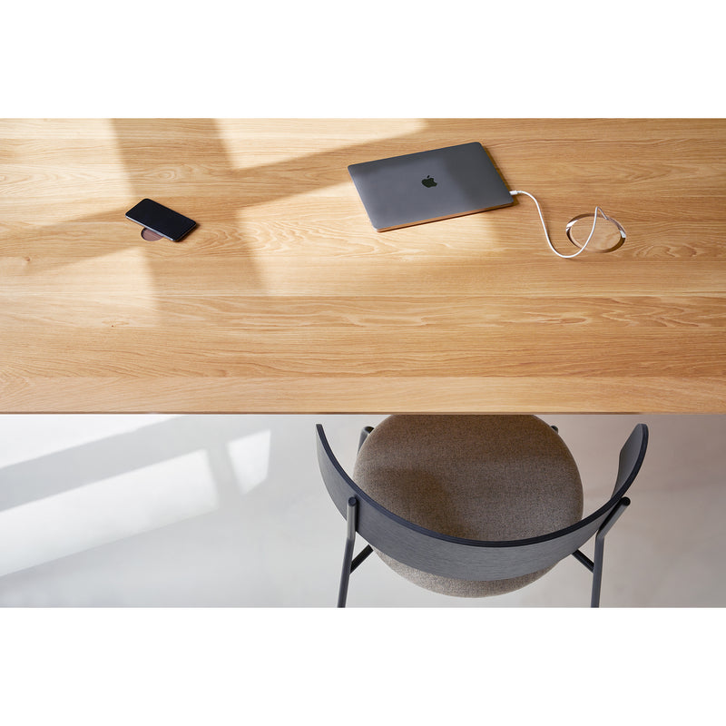 A Wedekind Table by Objekte unserer Tage, crafted from sustainable oak, holds a smartphone and charging laptop. A gray chair with a curved backrest is visible at the bottom, as shadows and natural light dance across the scene.