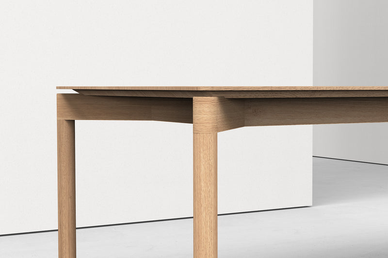 A minimalist wooden table sits against a plain white wall. The Wedekind Table by Objekte unserer Tage, crafted from sustainable oak, features a sleek finish and precise lines. A light gray floor enhances the scenes simplicity.