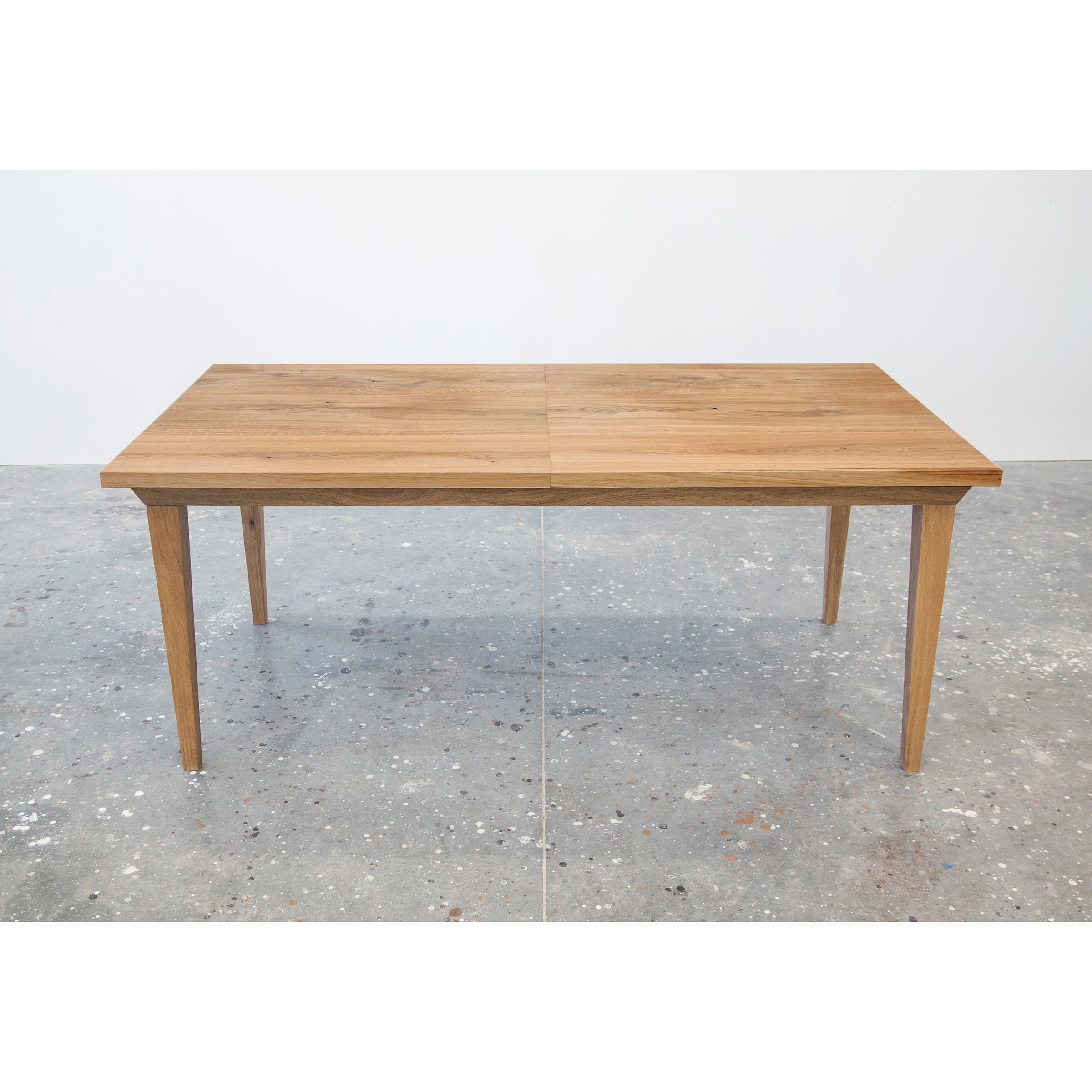 The Join Extendable Table from One For Hundred, crafted from solid wood with a natural finish, stands on a speckled concrete floor. This rectangular table features four tapered legs and an innovative synchronous extension mechanism for easy adjustments, maintaining a smooth minimalist design.