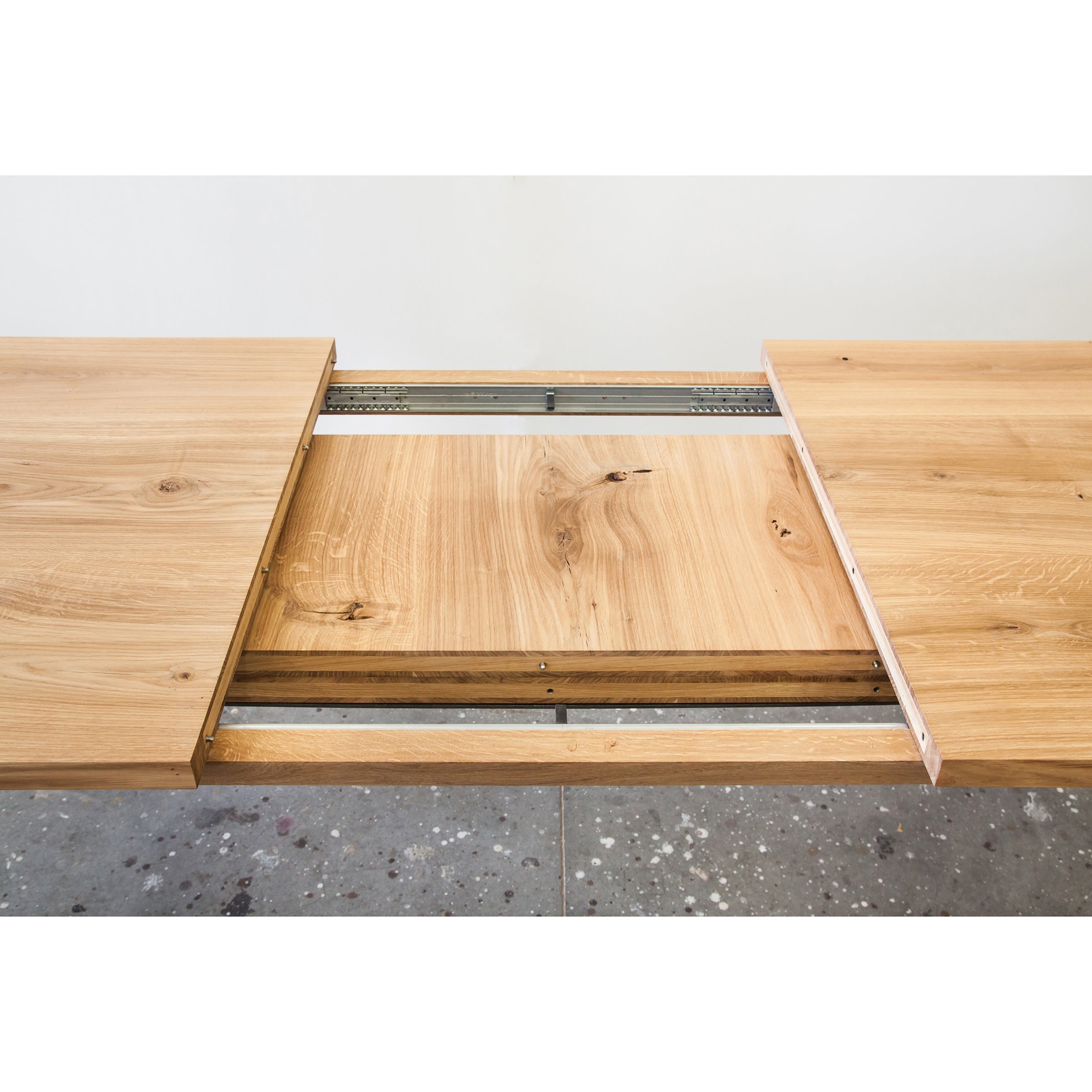 The Join Extendable Table by One For Hundred is beautifully crafted from solid wood. Its hidden extension leaf is partially revealed, showcasing the rich, light brown wood grain against a speckled gray floor.
