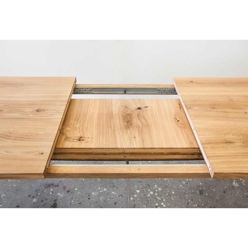 The Join Extendable Table by One For Hundred is beautifully crafted from solid wood. Its hidden extension leaf is partially revealed, showcasing the rich, light brown wood grain against a speckled gray floor.
