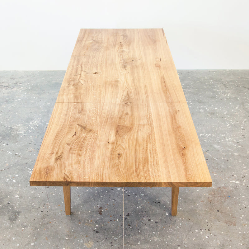 The Join Extendable Table by One For Hundred is a long, rectangular solid wood table with a natural grain finish, simple modern legs, and a polished surface. It stands on a character-speckled concrete floor and features an extendable design with a synchronous extension mechanism for added versatility.