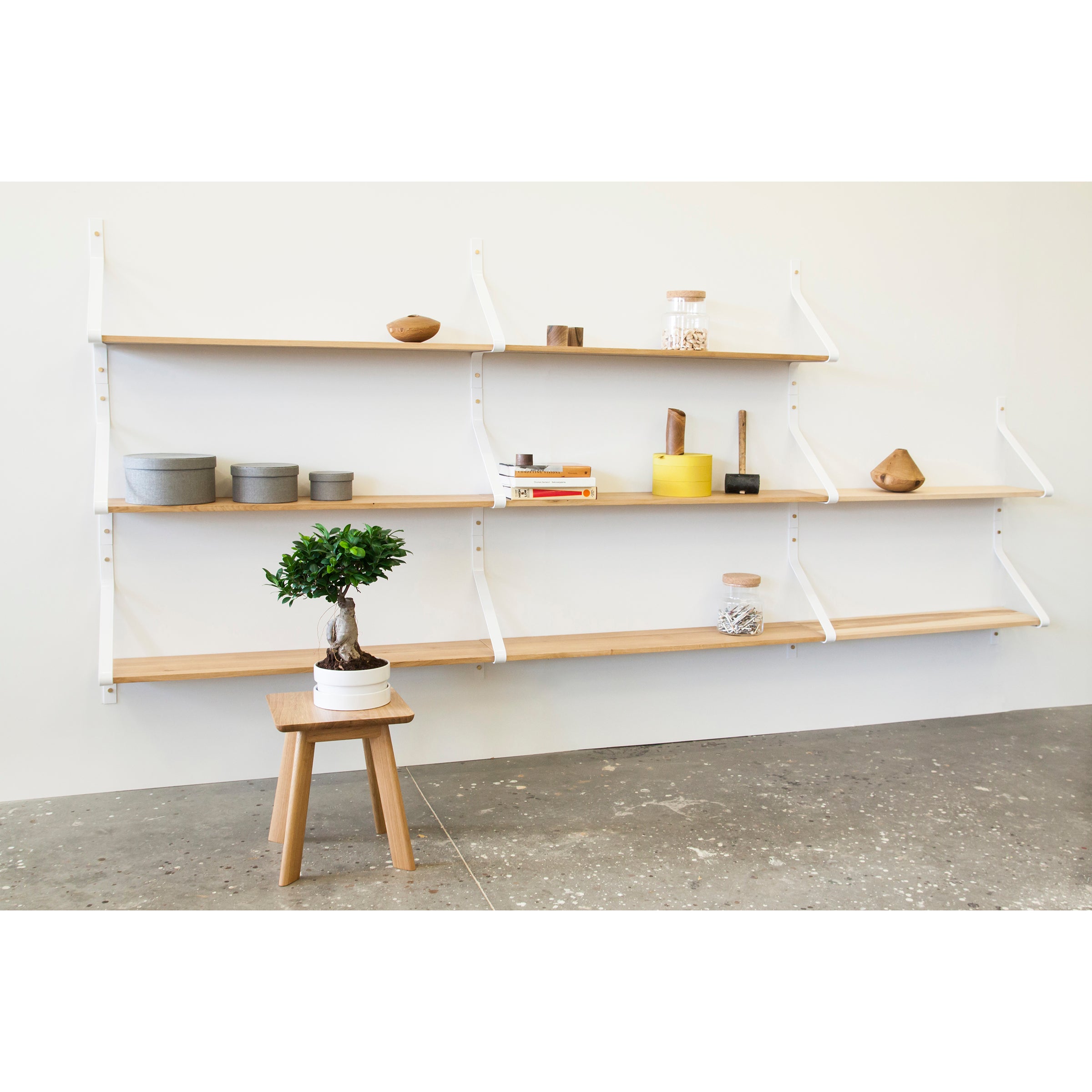 The Ribbon Shelf by One For Hundred, with its minimalist wooden design and steel brackets, showcases assorted pottery, a yellow container, books, and small decorations. A compact wooden stool below supports a bonsai tree in a white pot on the concrete floor.