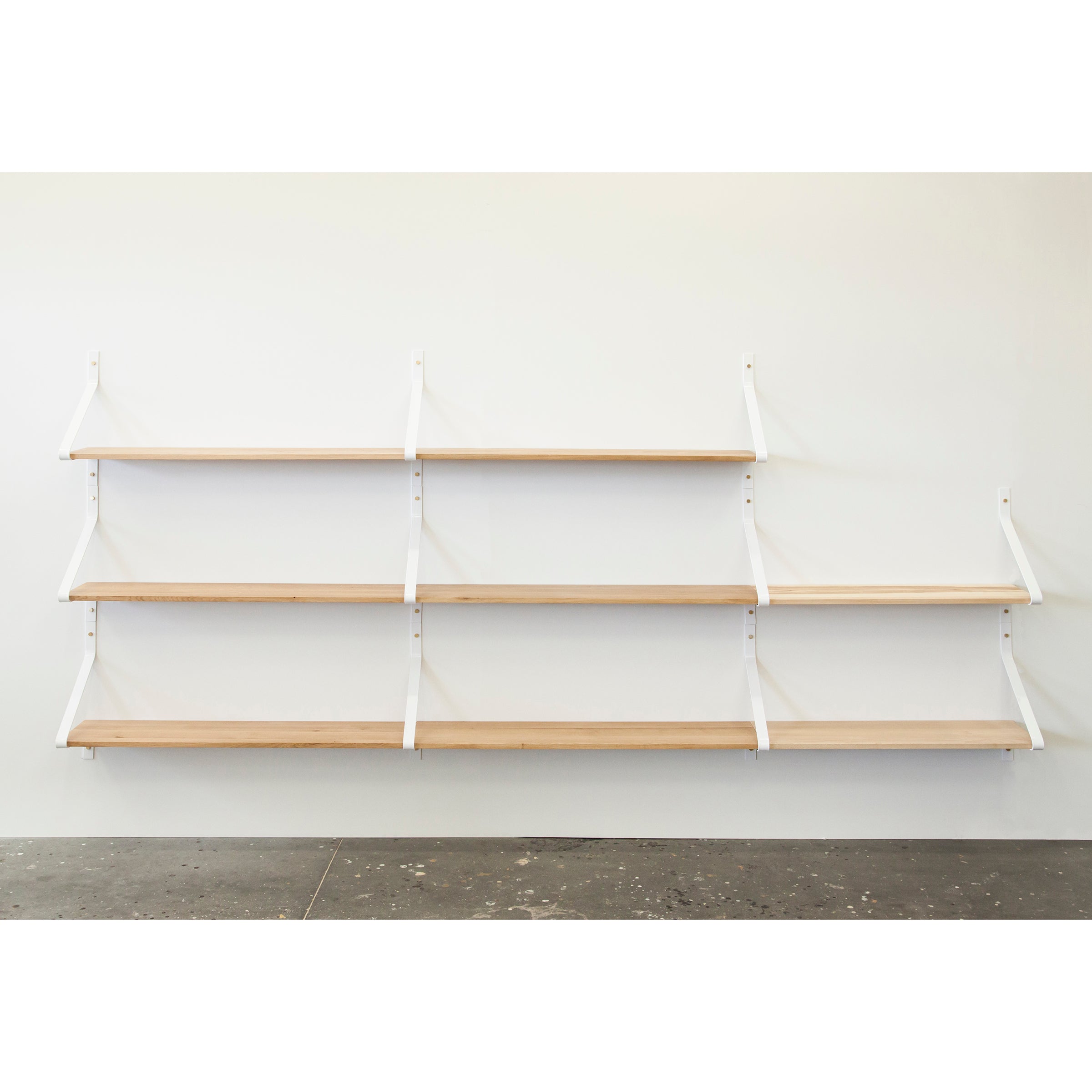 The Ribbon Shelf by One For Hundred is an elegant wall-mounted unit featuring four solid wood shelves, supported by white powder-coated steel brackets. Set against a plain white wall with a dark speckled floor, the evenly spaced shelves offer a sleek and graceful appearance.